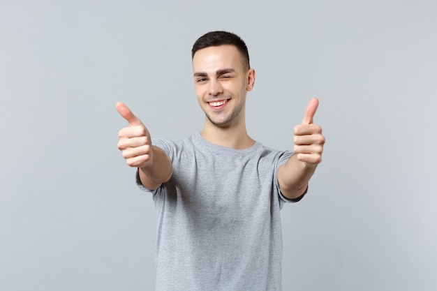 Portrait of cheerful blinking young man in casual clothes standing and showing thumbs up 