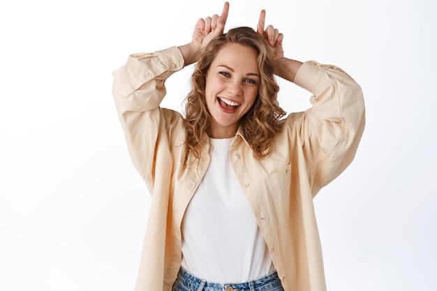 Portrait of cheerful blond girl stay positive, showing stubborn horns gesture on head, smiling and looking happy, standing over white background