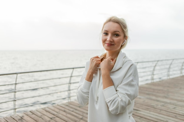 Portrait of a cheerful blonde sportswoman on the beach. Young slim fit woman listening to music with headphones