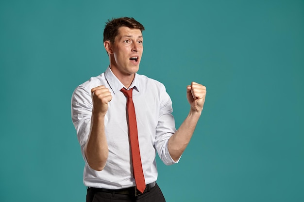 Portrait of a cheerful brunet fellow with brown eyes, wearing in a classic white shirt and red tie. He is acting like has won something posing in a studio against a blue background. Concept of gesticu