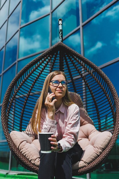 Portrait of a cheerful business woman in glasses enjoying a
coffee break outdoors on a sunny day a successful woman manager
talking on the phone while sitting against the background of the
office