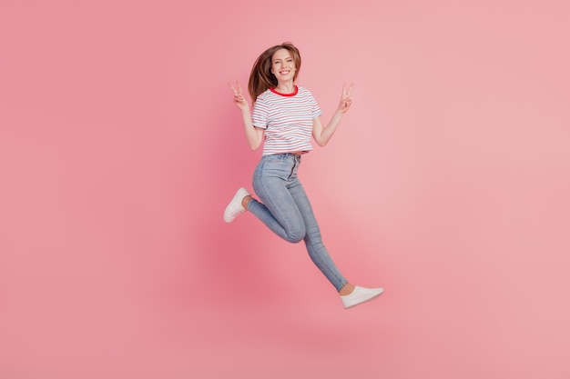 Portrait of cheerful carefree lady jump run have fun show v-signs on pink background