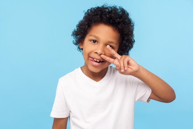 Portrait of cheerful carefree little boy with curly hair in Tshirt picking nose and sticking out tongue with happy face having fun bad manners concept studio shot isolated on blue background