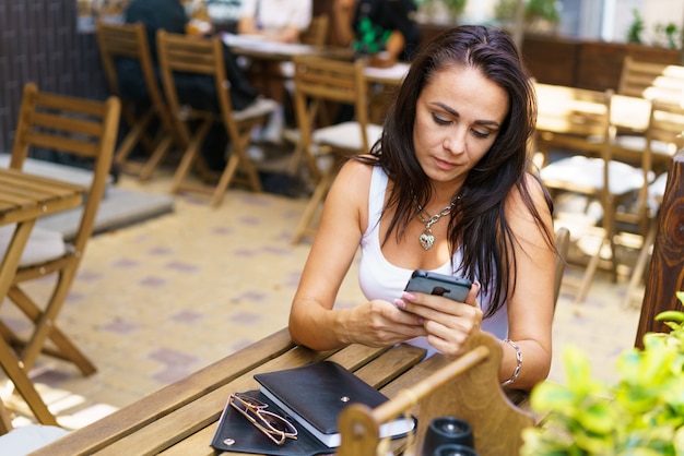 Portrait of cheerful caucasian woman looking at street while sitting in cafe with mobile phone and n...