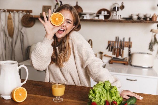 portrait of cheerful caucasian woman wearing casual clothes laughing and holding orange in cozy kitchen