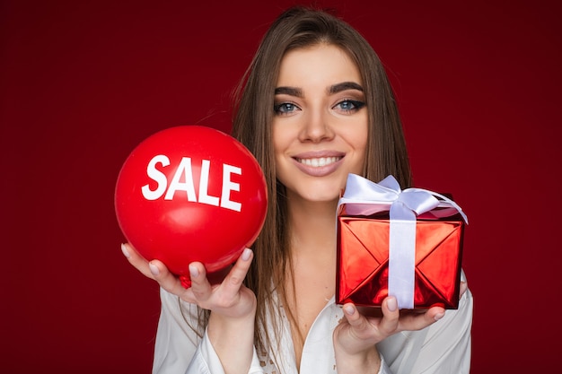 Portrait of cheerful caucasian woman with red baloon with sale on it on the one hand and red box with a gift on the other