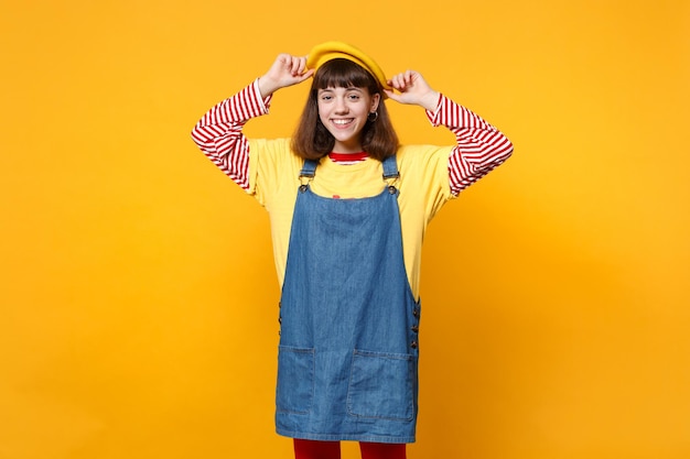 Portrait of cheerful charming girl teenager in denim sundress keeping hands on french beret isolated on yellow wall background in studio. People sincere emotions lifestyle concept. Mock up copy space.