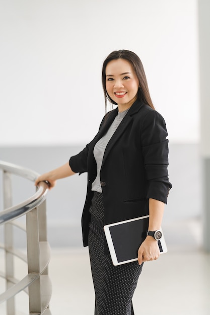 Portrait of a cheerful confident Asian businesswoman in a business suit standing whie using a digital tablet in the business building. Business stock photo