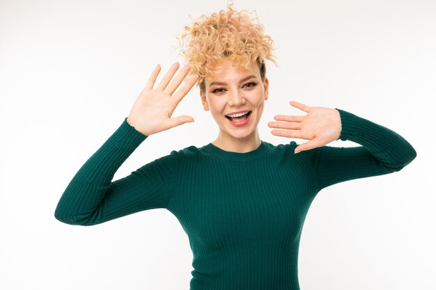 Portrait of a cheerful curly blond dressed in green clothes girl on a white wall