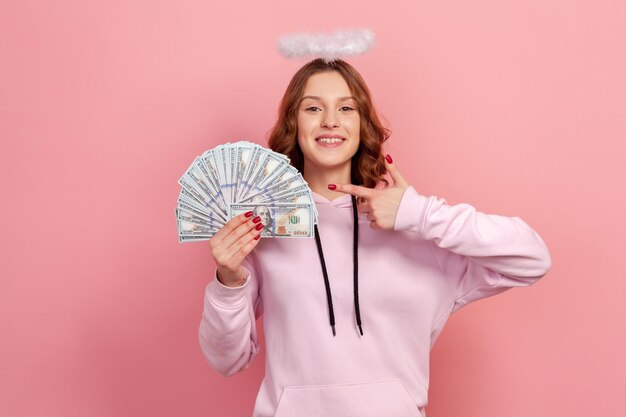 Portrait of cheerful curly haired teenage girl in hoodie with halo on head pointing finger on bunch of dollar banknotes and smiling profit Indoor studio shot isolated on pink background