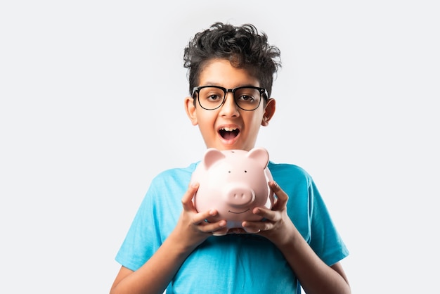 Portrait of a cheerful cute little Indian asian kid holding piggy bank with books  isolated over white wall