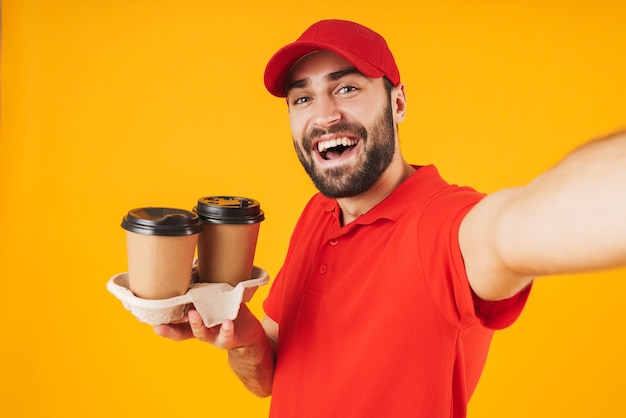 Portrait of cheerful delivery man in red uniform smiling and holding takeaway coffee cups while taking selfie photo isolated over yellow