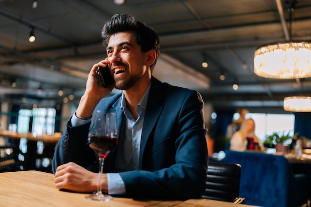 Portrait of cheerful elegant man wearing fashion suit talking on smartphone sitting at table with glass of red wine in restaurant