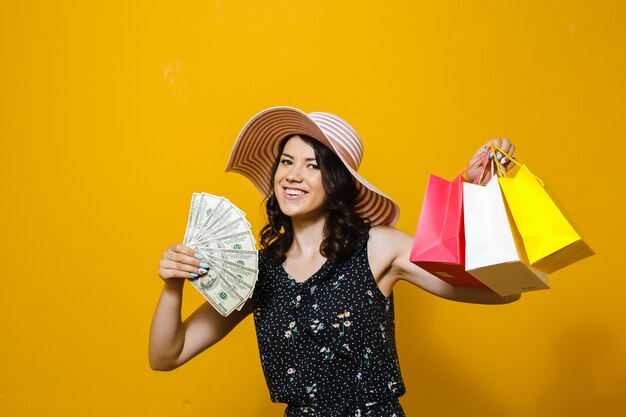 Portrait of cheerful elegant young woman in sunglasses and hat holding  money and colorful shopping bags isolated over yellow background