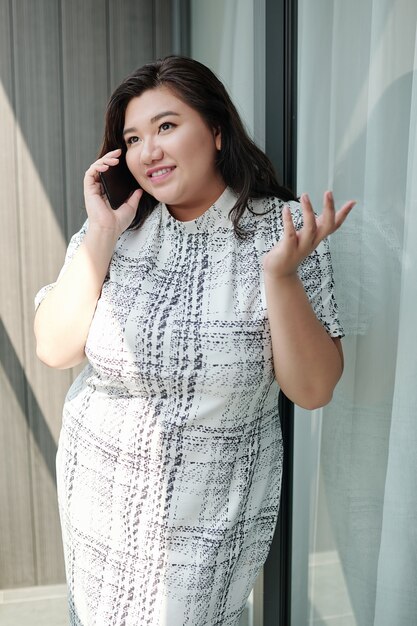 Portrait of cheerful emotional young plus size woman standing on balcony and talking on phone