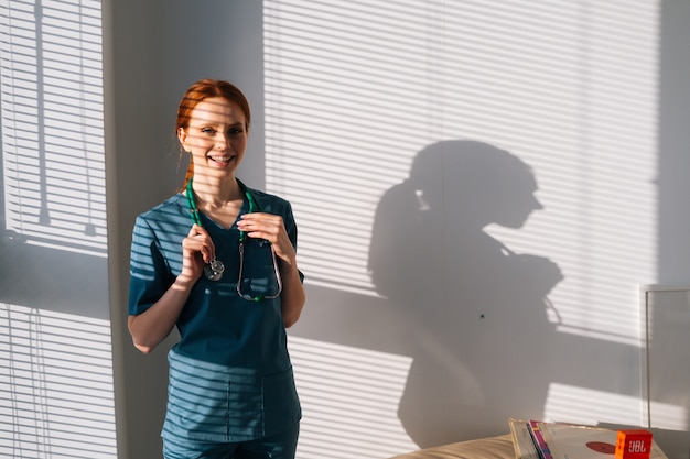 Portrait of cheerful female doctor in blue green uniform standing near window in sunny day in medical clinic office.