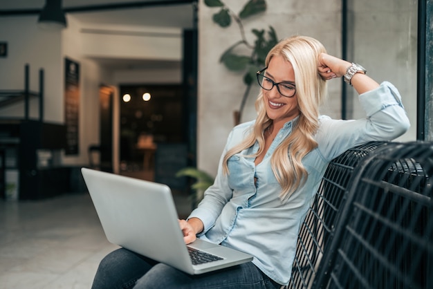 Photo portrait of cheerful female freelancer in eyewear using laptop indoors.