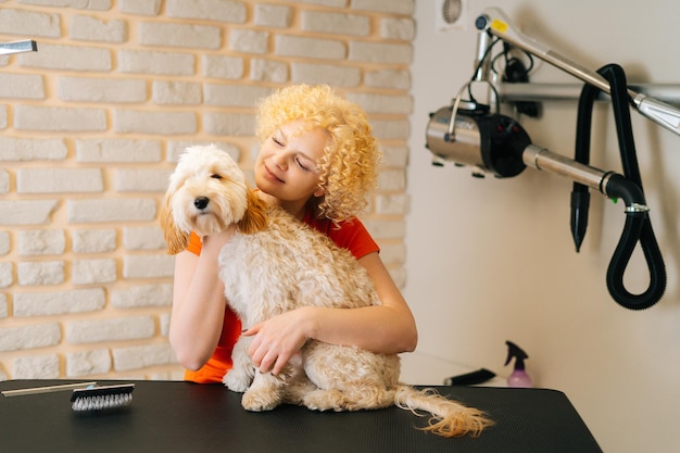 Portrait of cheerful female groomer hugging adorable curly\
labradoodle dog before brushing and shearing, preparation to\
bathing at grooming salon. woman pet hairdresser doing professional\
care.