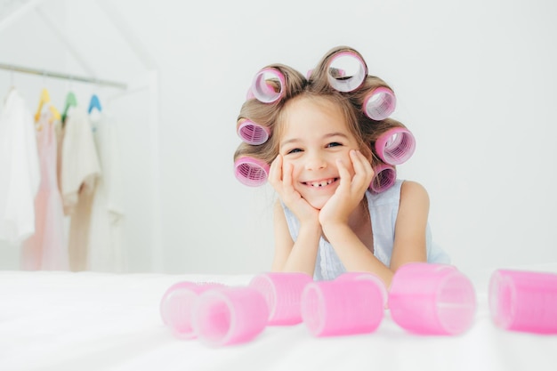 Portrait of cheerful female kid keeps hands under chin has curlers on hair going to have nice hairstyle poses against white background has charming smile being in good mood Children and beauty