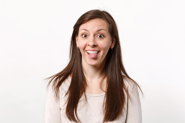 Portrait of cheerful funny crazy young woman in light clothes looking camera showing tongue isolated on white wall background in studio. People sincere emotions, lifestyle concept. Mock up copy space.