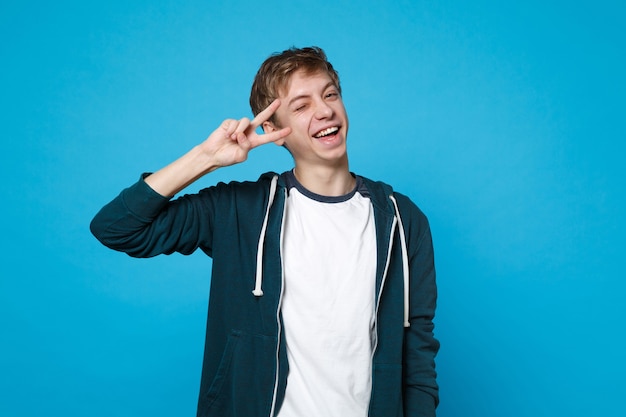 Portrait of cheerful funny young man in casual clothes blinking and showing victory sign isolated on blue wall. 