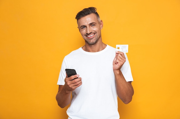 黄色で隔離の携帯電話とクレジットカードを保持している白いTシャツの陽気な男の30代の肖像画