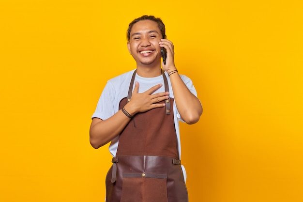 Portrait of cheerful handsome Asian young man wearing apron talking on mobile and hands on chest