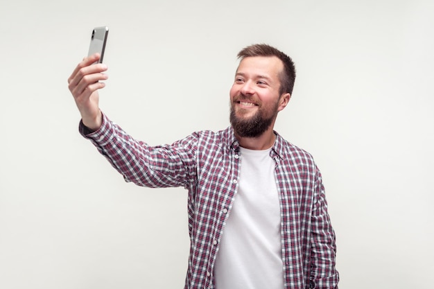 Portrait of cheerful handsome bearded man in casual plaid shirt taking selfie using smartphone, talking on video call and smiling joyfully, mobile communication. indoor studio shot, white background