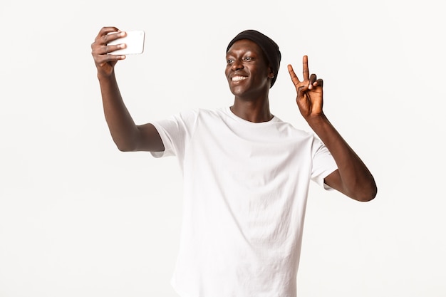 Portrait of cheerful handsome Black guy in beanie, taking selfie on mobile phone and showing peace gesture happy, smiling