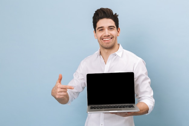 Portrait of a cheerful handsome young man standing isolated on blue, working on laptop computer, showing blank screen laptop