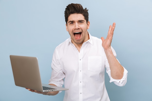 Portrait of a cheerful handsome young man standing isolated on blue, working on laptop computer