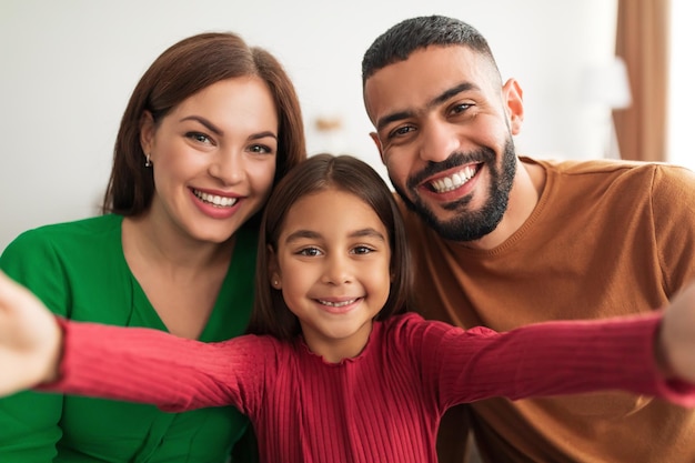 Portrait of cheerful happy family taking selfie together at home indoors. Happy parents posing with their child daughter and smiling at camera, point of view pov. Bonding And Lifestyle Concept.