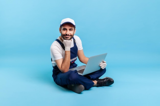 Portrait of cheerful happy professional handyman in workwear sitting cross-legged with laptop, leaning on hand and looking at camera with friendly smile. indoor studio shot isolated on blue background