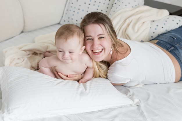 Portrait of cheerful laughing mother relaxing on bed with her 9 months old baby boy