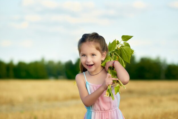 Portrait of a cheerful little cute little girl in a summer dress with a bouquet in her hands walking along a field outside the city on a warm summer evening