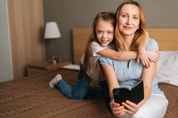 Portrait of cheerful little girl hugging to beautiful mother holding mobile phone and looking at camera, chatting on video call sitting on bed in bedroom. Concept of family leisure activity at home
