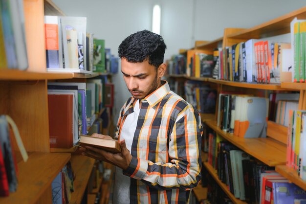 Portrait of cheerful male international Indian student with backpack learning accessories standing near bookshelves at university library or book store during break between lessons Education concept