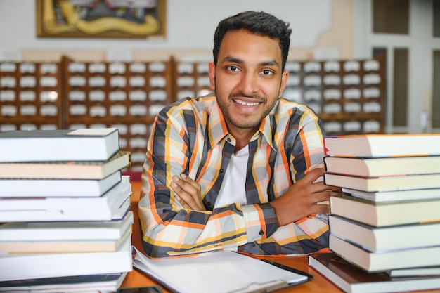 レッスン間の休憩中に大学図書館や本屋の本棚の近くに立っているバックパック学習アクセサリーを持つ陽気な男性国際インド学生のポートレート教育コンセプト