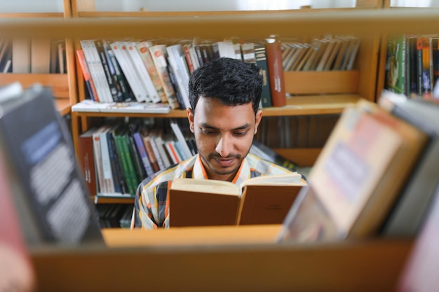 レッスン間の休憩中に大学図書館や本屋の本棚の近くに立っているバックパック学習アクセサリーを持つ陽気な男性国際インド学生のポートレート教育コンセプト