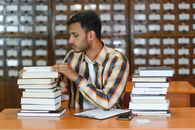 Portrait of cheerful male international Indian student with backpack learning accessories standing near bookshelves at university library or book store during break between lessons Education concept