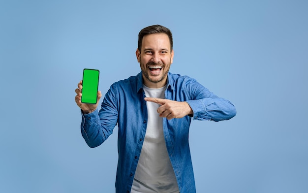 Portrait of cheerful male professional pointing at green smart phone screen against blue background