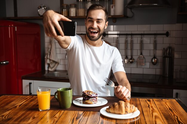 Portrait of cheerful man 30s taking selfie photo on mobile phone while having breakfast in stylish kitchen at home