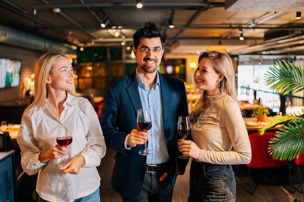 Портрет веселого мужчины и двух подруг-блондинок, держащих бокалы красного вина, улыбающихся друг другу, стоящих и позирующих в ресторане