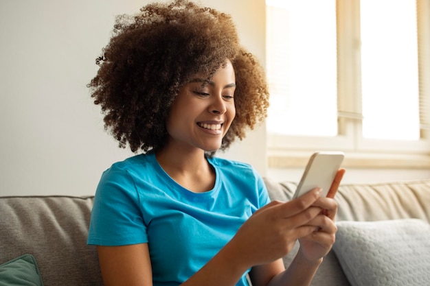 Portrait of cheerful millennial curly black lady typing on smartphone read message watch video