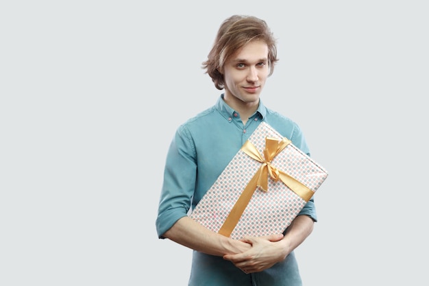 Portrait of cheerful modern young man in light blue shirt standing and holding a present with yellow bow, looking at camera and smile. Indoor, isolated, studio shot, grey background