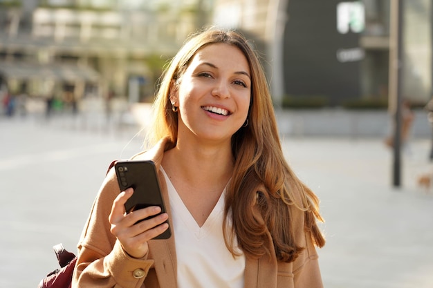 Portrait of cheerful multi ethnic female in trendy wear spending time on street using smartphone, beautiful millennial girl with coat looking at camera in town holding mobile phone