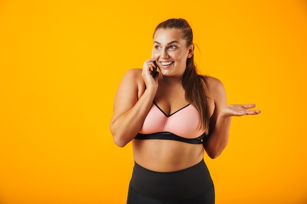 Portrait of a cheerful overweight fitness woman wearing sports clothing standing isolated over yellow wall, talking on mobile phone