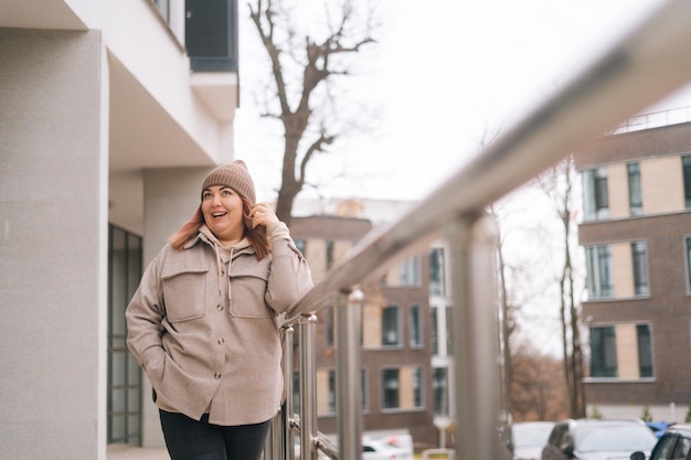 Portrait of cheerful overweight woman in warm hat and jacket standing posing near railing of office building at city street in cloudy autumn day