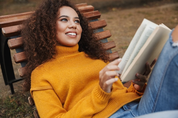 Portrait of cheerful pleased beautiful curly woman lies in park outdoors reading book.