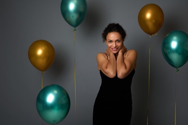Portrait of cheerful pretty woman in evening black dress smiling with beautiful toothy smile posing against air balloons on gray wall background with copy space for Christmas, New Year, anniversary ad